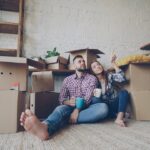 Couple sitting on a rug among moving cardboard boxes in a plant-filled room, the man holding a blue mug as the woman points at something off to the side.