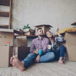 Couple sitting on a rug among moving cardboard boxes in a plant-filled room, the man holding a blue mug as the woman points at something off to the side.